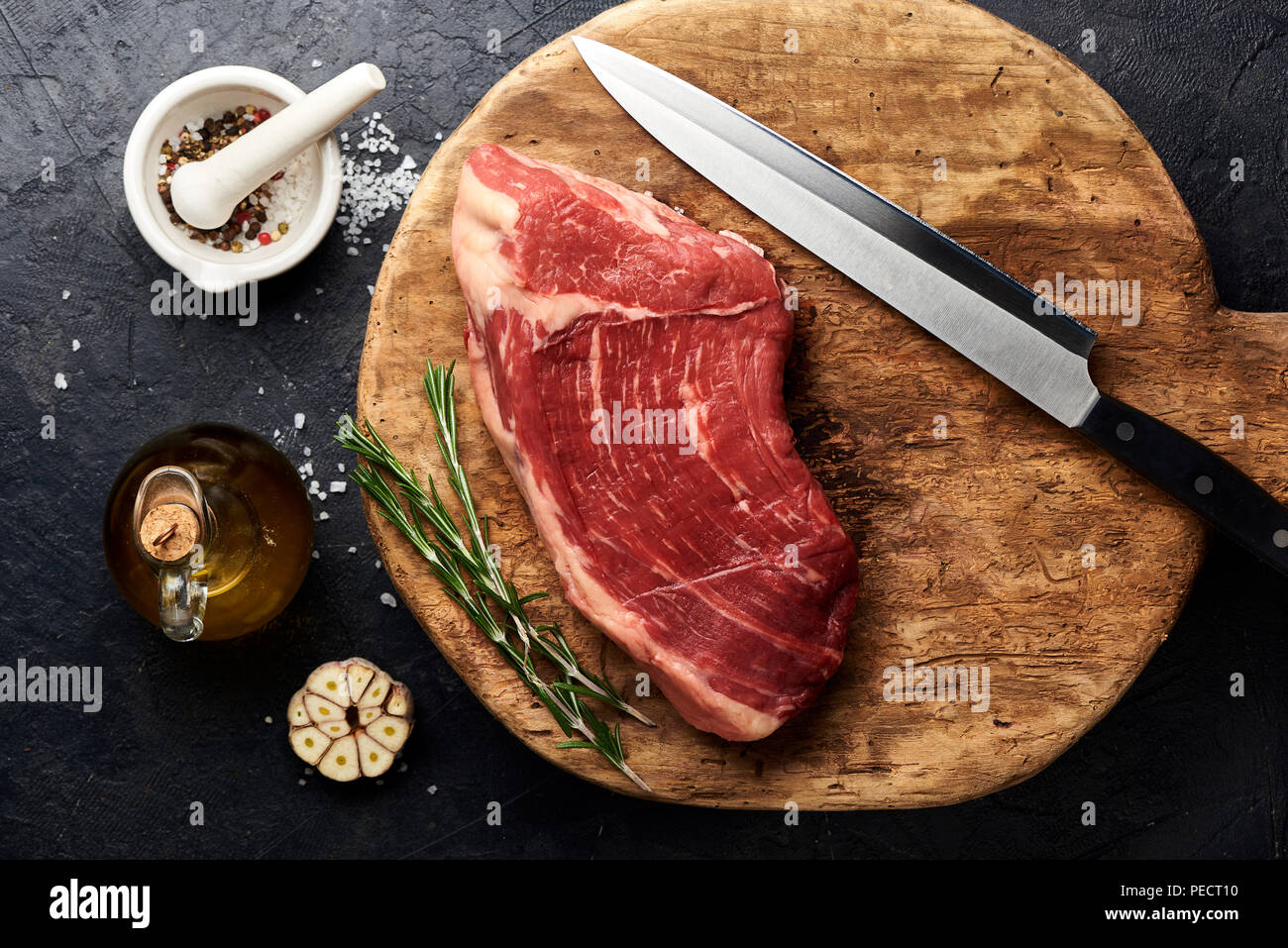 Rohe frische marmoriertes Fleisch Black Angus Steak mit Messer für Fleisch auf Holzbrett. Fleisch auf schwarzen Hintergrund mit Rosmarin, Gewürze, Olivenöl und Knoblauch. Kopieren Sie Platz. Ansicht von oben. Stockfoto