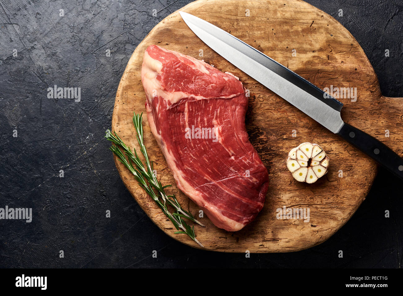 Rohe frische marmoriertes Fleisch Black Angus Steak mit Messer für Fleisch auf Holzbrett. Fleisch auf schwarzen Hintergrund mit Rosmarin und Knoblauch. Kopieren Sie Platz. Ansicht von oben. Stockfoto