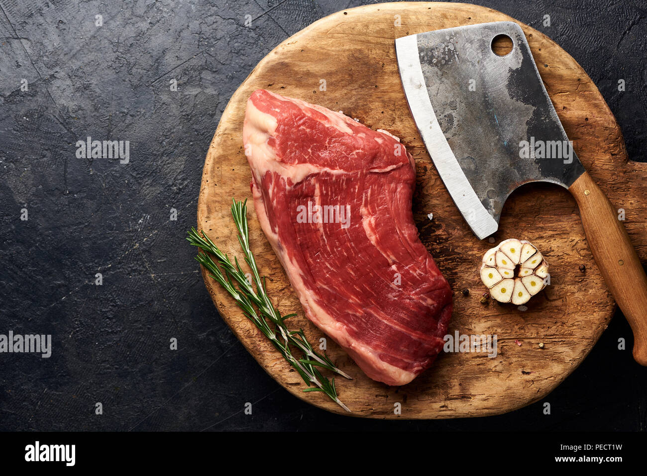 Rohe frische marmoriertes Fleisch Black Angus Steak und Fleisch Cleaver auf Holzbrett. Fleisch auf schwarzen Hintergrund mit Rosmarin und Knoblauch. Kopieren Sie Platz. Ansicht von oben. Stockfoto