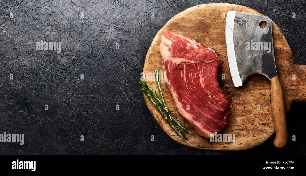 Rohe frische marmoriertes Fleisch Black Angus Steak und Fleisch Cleaver auf Holzbrett. Fleisch auf schwarzen Hintergrund mit Rosmarin. Kopieren Sie Platz. Ansicht von oben. Stockfoto