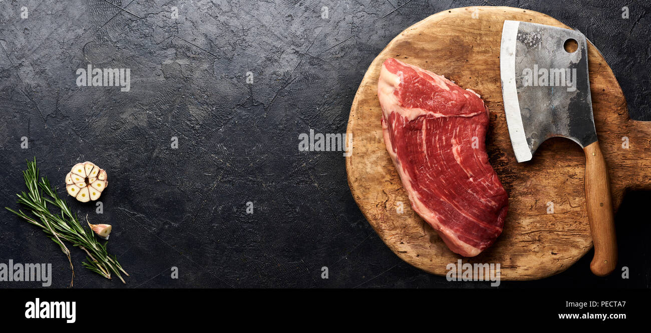 Rohe frische marmoriertes Fleisch Black Angus Steak und Fleisch Cleaver auf Holzbrett. Fleisch auf schwarzen Hintergrund mit rosemarym Knoblauch. Kopieren Sie Platz. Ansicht von oben. Stockfoto