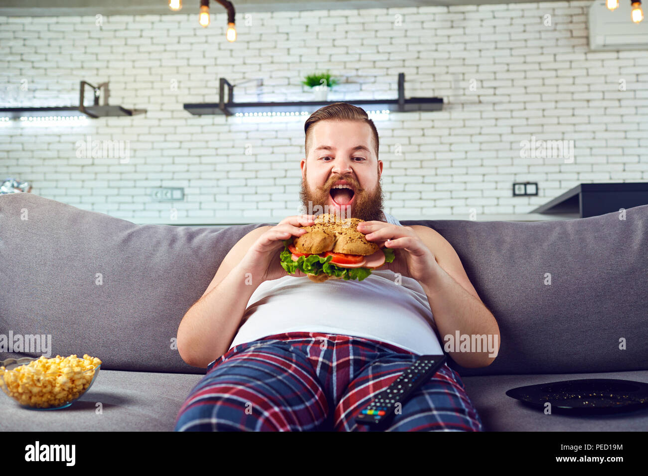 Fett lustig Mann im Schlafanzug essen einen Burger auf dem Sofa zu Hause. Stockfoto