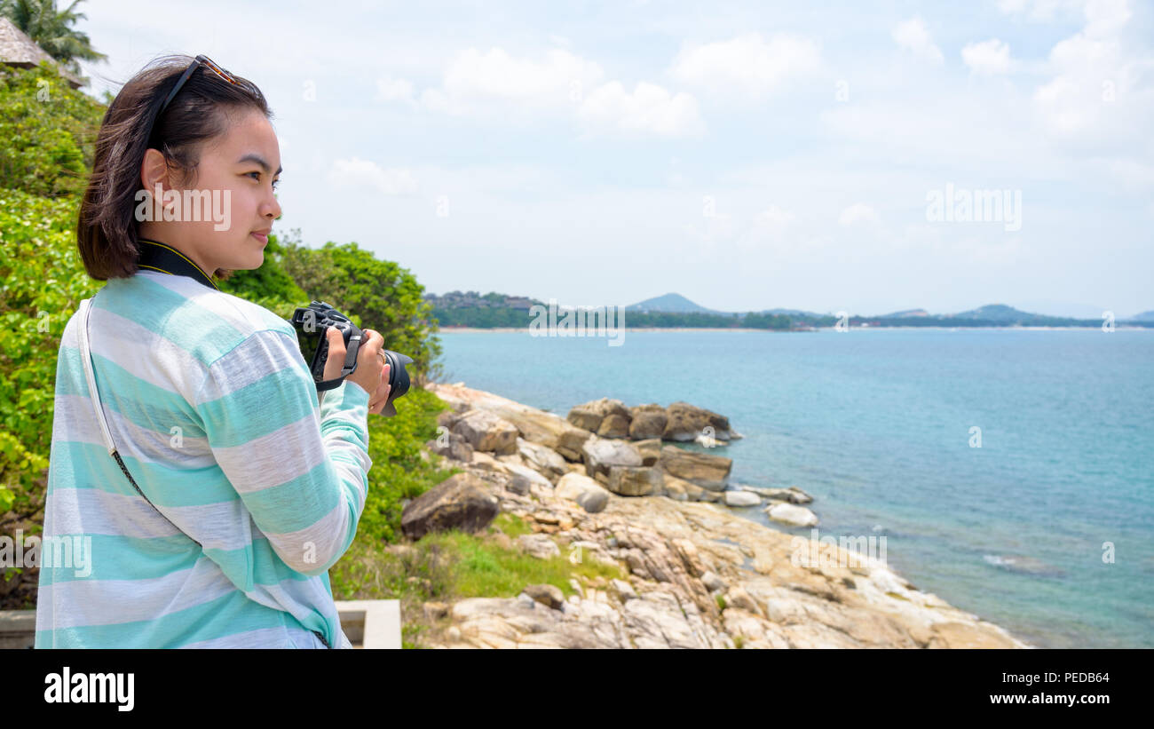 Junge Frau glücklich war mit DSLR-Kamera, die auf dem Felsen in der Nähe des Meeres zu Fotografie unter dem Sommer Himmel in Koh Samui, Surat Thani Provinz, Thailan Stockfoto