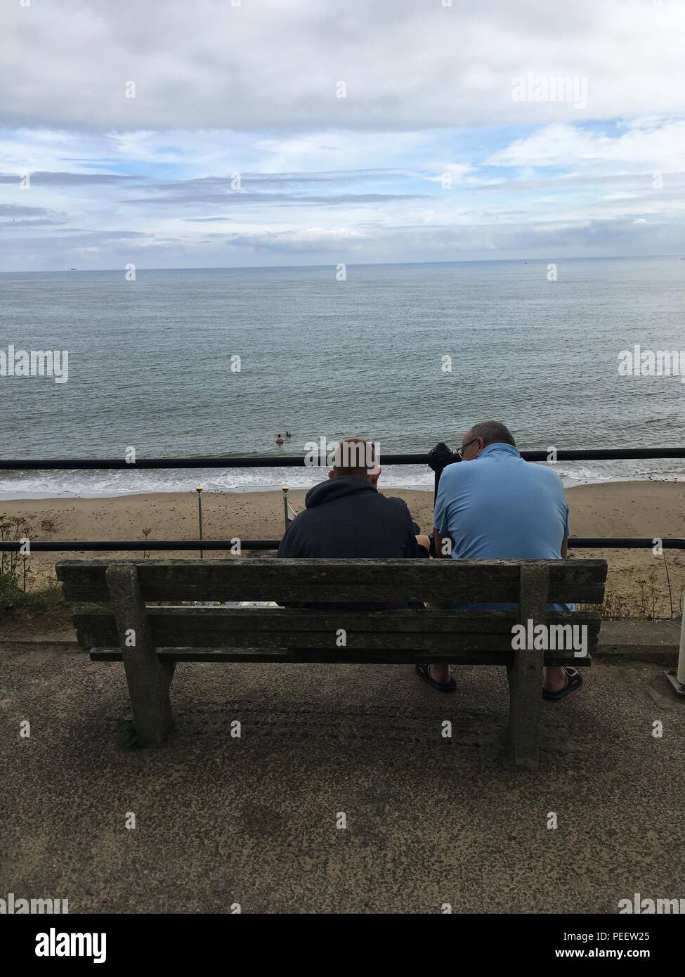 Erwachsene männliche und junge Männer sitzen auf einer Bank mit Blick auf das Meer Stockfoto