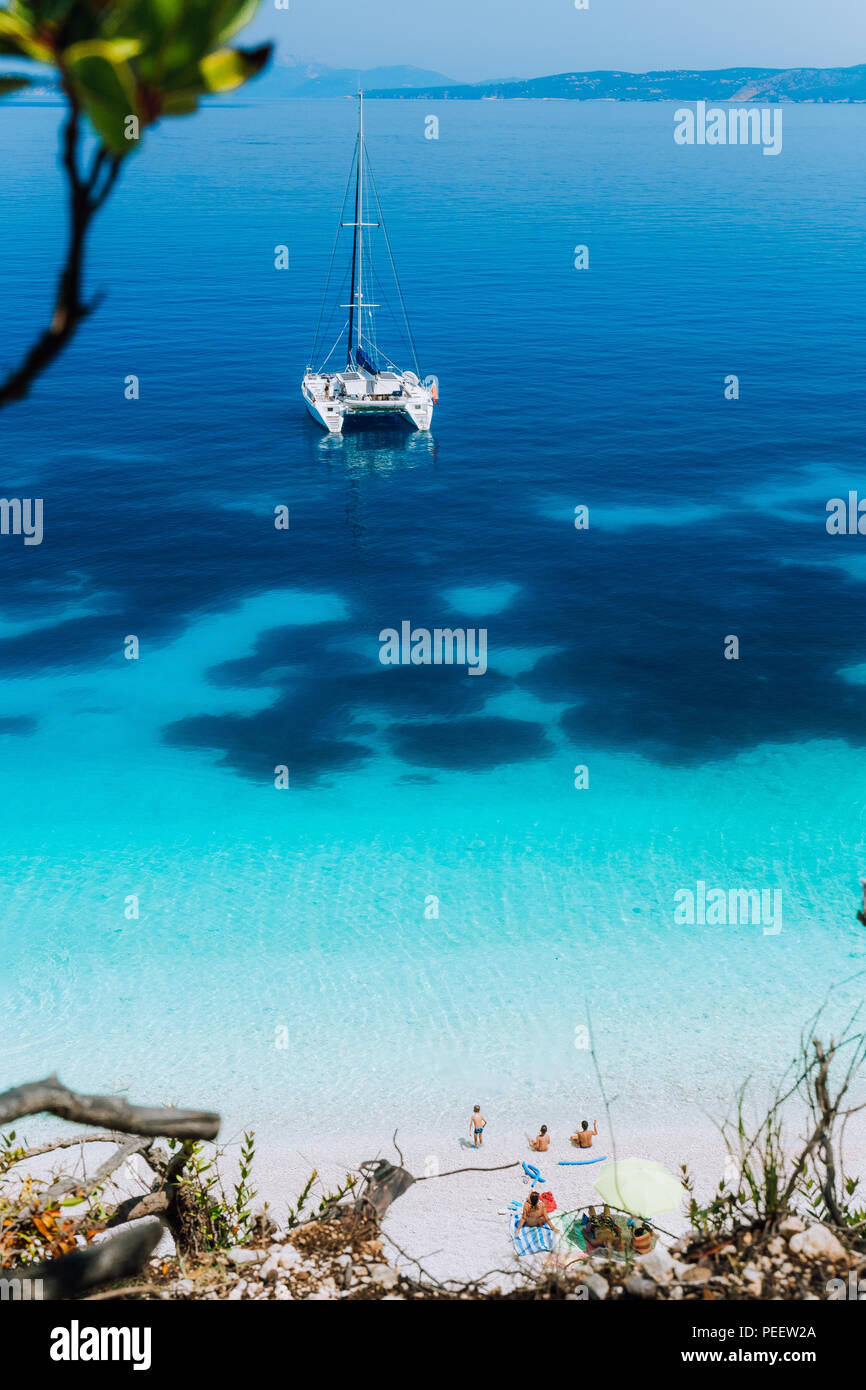 Weißen Katamaran Yacht am azurblauen Oberfläche Anker mit dunklem in ruhigen blauen Lagune. Nicht erkennbare Touristen unter Sonnenschirm am Strand entspannen Stockfoto