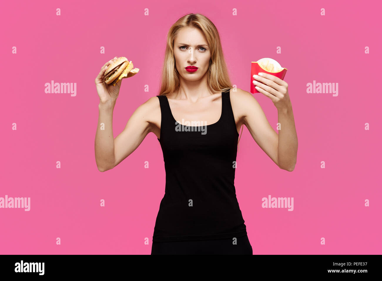 Junge schöne schlanke Mädchen auf einem rosa Hintergrund hält einen Burger und Pommes frites in den Händen. Konzept der "junk food" Stockfoto