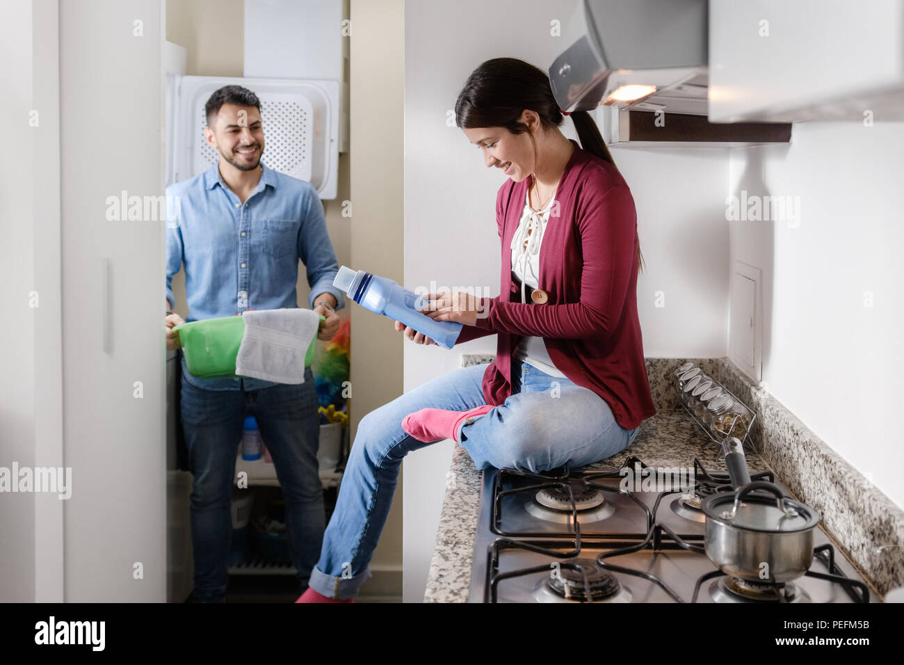 Junges Paar, Hausarbeit und Hausarbeiten. Man Kleidung, innen Waschmaschine und Freundin sitzen auf den Küchentisch, lesen Label auf reinigt Stockfoto