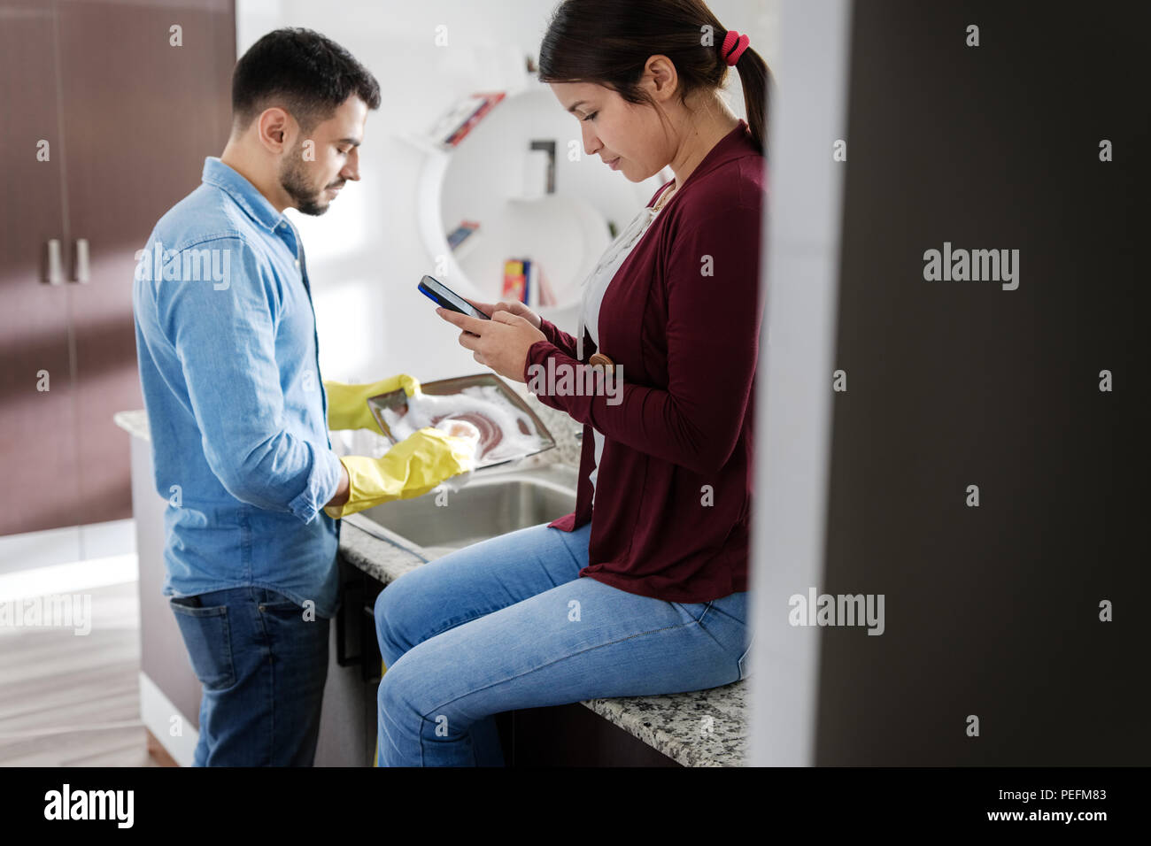 Junges Paar, Hausarbeit und Hausarbeiten. Mann Reinigung Geschirr in der Küche und der Freundin sitzen auf Zähler, Handy und lacht. Stockfoto