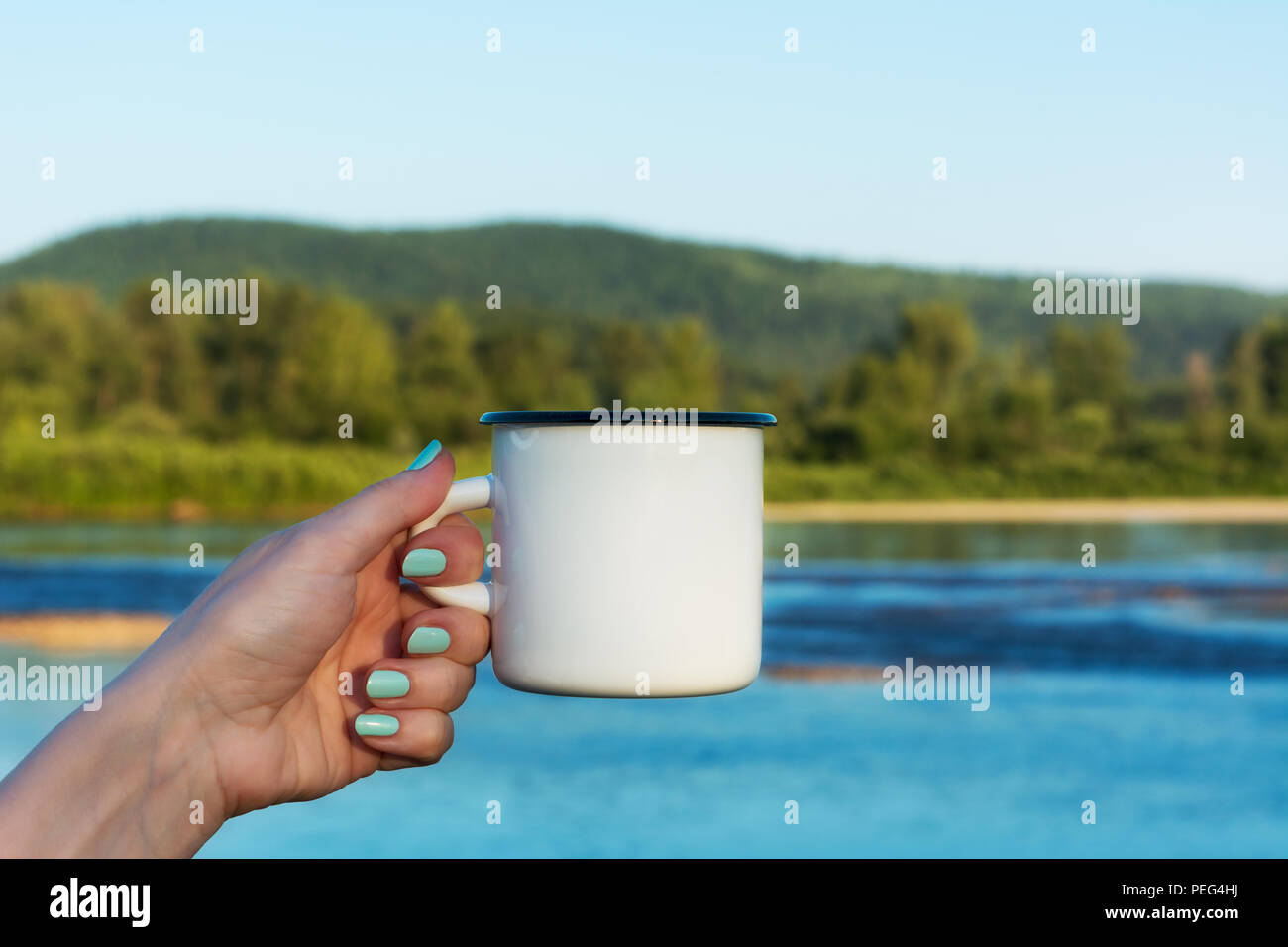 Frau mit einem weißen Lagerfeuer Emaille Tasse mockup mit Sommerterrasse mit Blick auf den Fluss. Leere Becher mock up für Design Promotion. Stockfoto