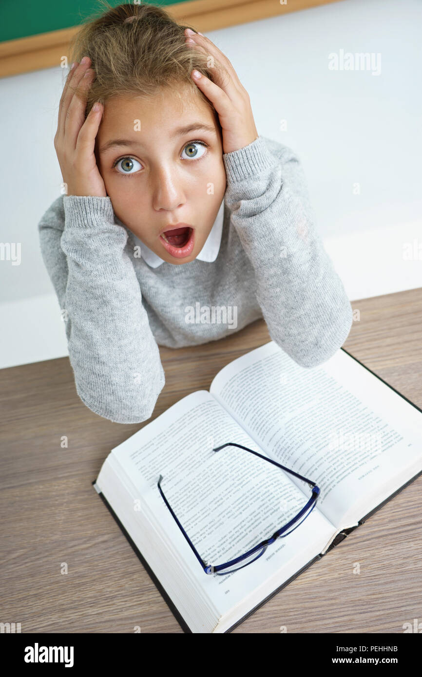 Kleines Schulmädchen holding Kopf mit geöffnetem Mund und verrückten Gesichtsausdruck. Foto von Mädchen mit Buch auf dem Schreibtisch im Klassenzimmer. Zurück zur Schule Stockfoto