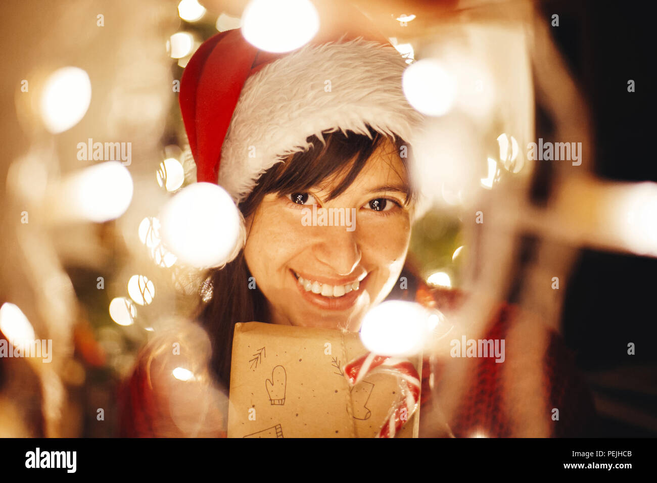 Frohe Weihnachten. Happy girl in Santa Hat und Red Pullover holding Weihnachten in Lichter am Abend festliches Zimmer im Baum Beleuchtung vorhanden. Öffnen des mod Stockfoto