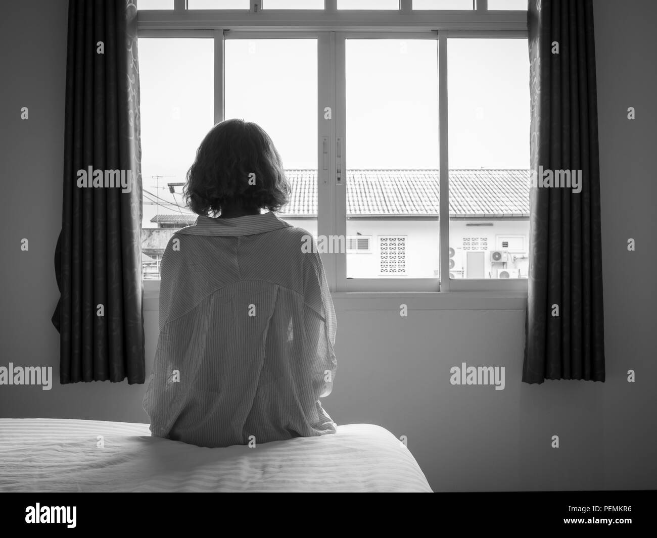 Frau kurze Haare sitzen auf dem weißen Bett im Hotelzimmer. Einsame Frau, die man vom Fenster aus sieht allein in Weiß, Schwarz und Weiß. Stockfoto
