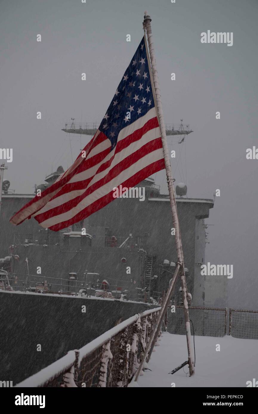 Am 24. Januar 2016, an Bord der USS Ashland (LSD 48) in Sasebo, Japan, flattert die Fähnrich auf dem schneebedeckten Flugdeck, bevor die Abendfarben erscheinen. Das amphibische Dock-Landungsschiff, Teil der Bonhomme Richard Amphibious Ready Group, ist dabei, die geplante Continuous Maintenance Availability (CMAV) abzuschließen. Die Seeleute sind aufmerksam, während die Zeremonie den Respekt für das nationale Fähnrich und die Einsatzbereitschaft bei routinemäßigen Instandhaltungs- und Ausbildungseinsätzen kennzeichnet. Stockfoto