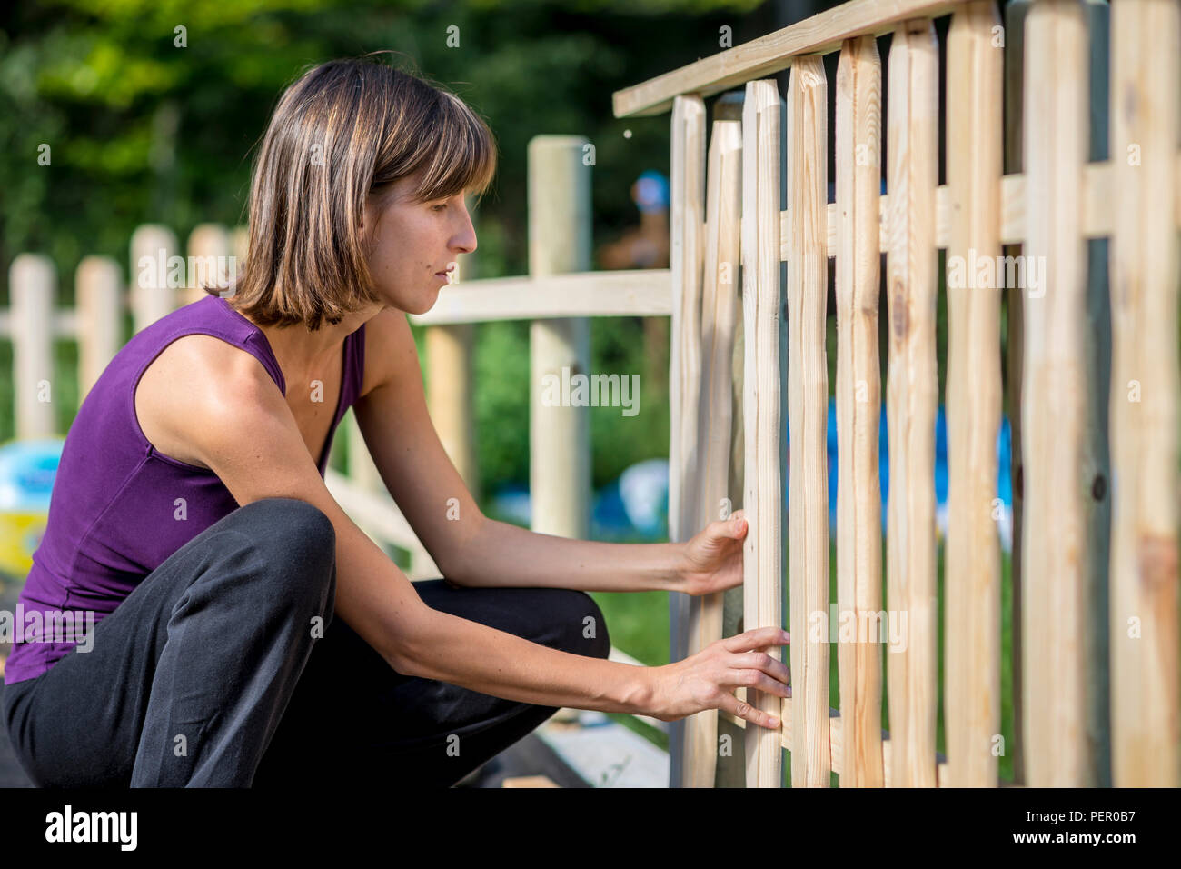 Frau Gebäude einen Gartenzaun eine aufrechte Holzstäbchen in Position in der Nähe zu seitlichen Blick auf ihren Knien zu ausgearbeitet werden. Stockfoto