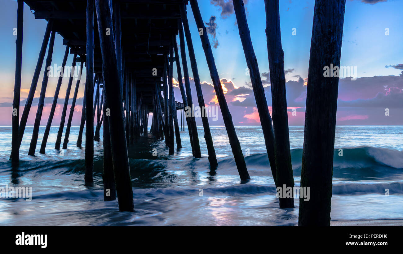 Kleine Wellen brechen gegen die hölzerne Stützen für ein Meer Pier. Sonnenaufgang am Horizont macht ein farbenfroher und dramatischer Himmel. Stockfoto