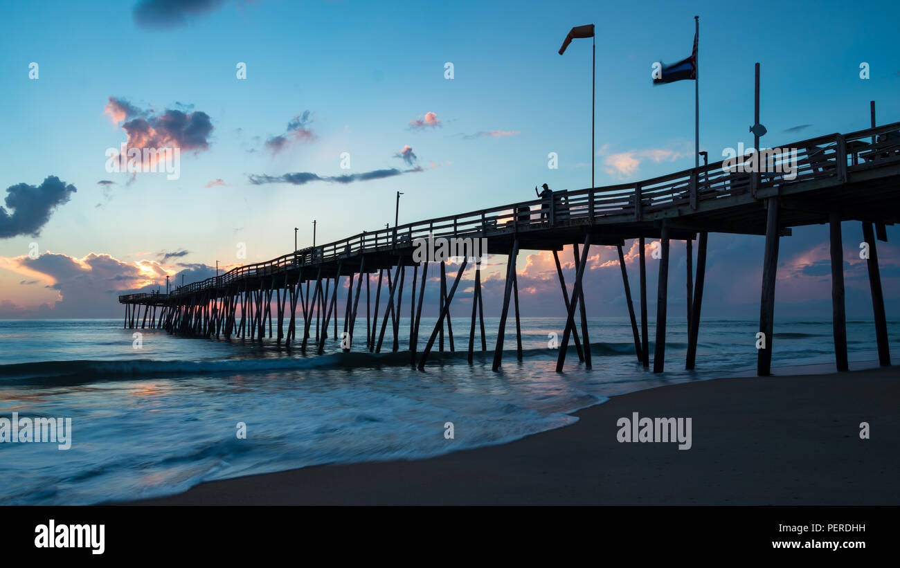 Eine lange hölzerne ocean Pier erstreckt sich in das Meer zu einem genialen Sonnenaufgang wie Fahnen im Wind fliegen. Stockfoto