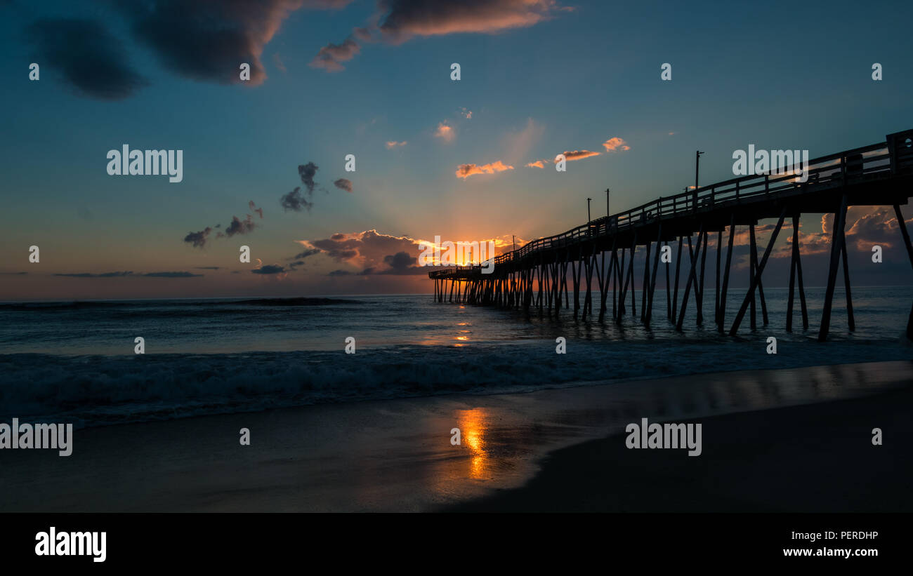 Lichtstrahlen, die platzen aus den Wolken wie Sunrise Lichter das Meer Horizont am Ende einer Fishing Pier. Stockfoto