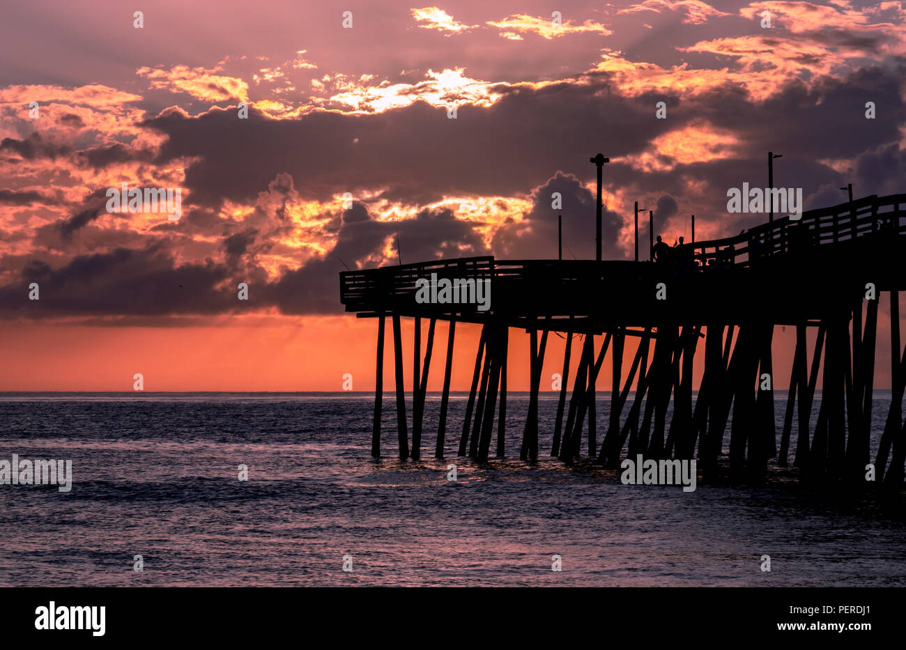 Sunrise leuchtet der Himmel mit rot und orange Licht hinter weichen Wolken. Eine ocean Fishing Pier im ersten Licht des Tages mit einem ruhigen Meer. Stockfoto