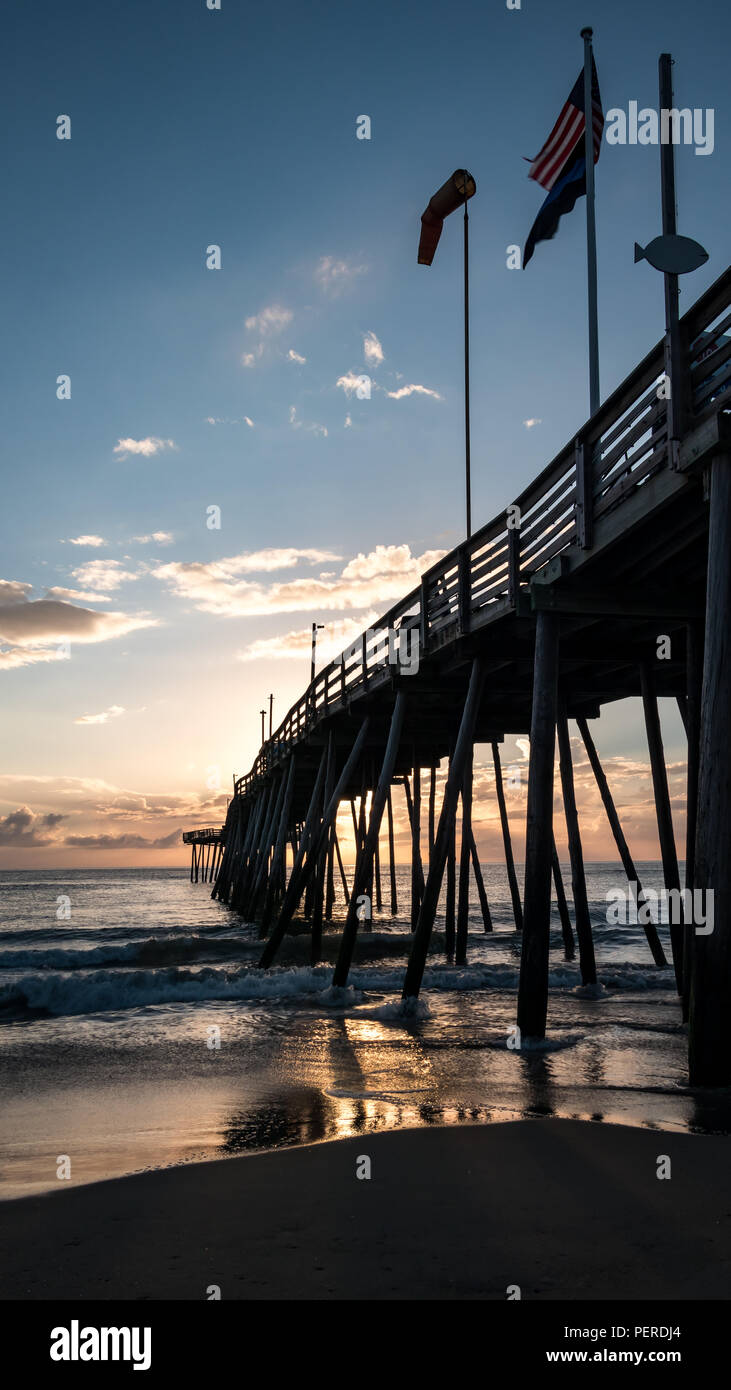 Fahnen wehen auf einer hölzernen Fishing Pier, die weit ins Meer erstreckt. Sunrise Farben in der Luft und im Wasser und auf den Sand nieder. Stockfoto