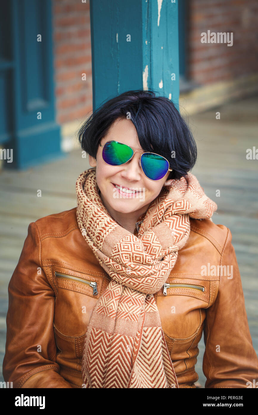 Portrait von lächelnden mittleren Alters kaukasischen Dunkelhaarige brünette Frau mit kurzen Bob Haar in Sonnenbrille trägt Hellbraun leder jacke und Schal o Stockfoto