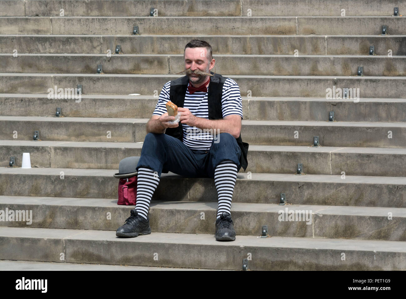Freudiger Mann mittleren Alters in gestreifter Clown Kostüm sitzt auf der Treppe der Vorbereitung einer sandvich zu essen Stockfoto