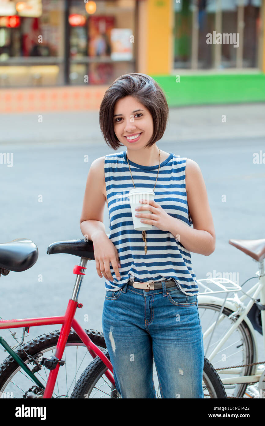 Porträt der Schönen lächelnden Jungen latin kolumbianische Mädchen Frau mit kurzen Haaren Bob in Blau zerrissene Jeans gestreiftes T-Shirt, Tasse Kaffee schiefen o Stockfoto