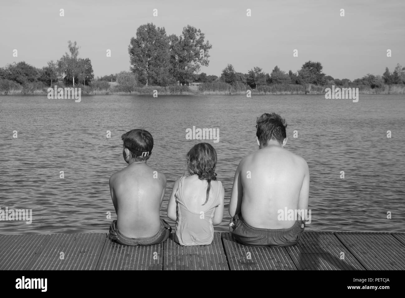 Kinder sitzen auf Pier. Drei Kinder verschiedenen Alters - jugendlicher Junge, elementare alter Junge und Mädchen sitzen auf einem hölzernen Pier. Sommer und Kindheit Konzept. Kinder auf der Bank am See. Geschwister sitzen auf hölzernen Pier Stockfoto