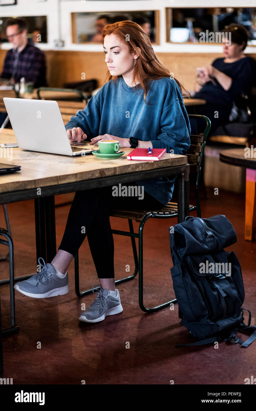 Junge Frau mit langen roten Haaren sitzen am Tisch, Arbeiten am Laptop. Stockfoto