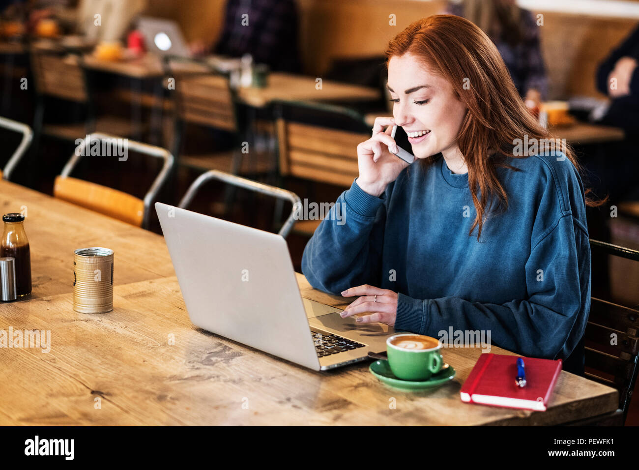 Lächelnde junge Frau mit langen roten Haaren sitzen am Tisch, Arbeiten am Laptop, mit Handy. Stockfoto