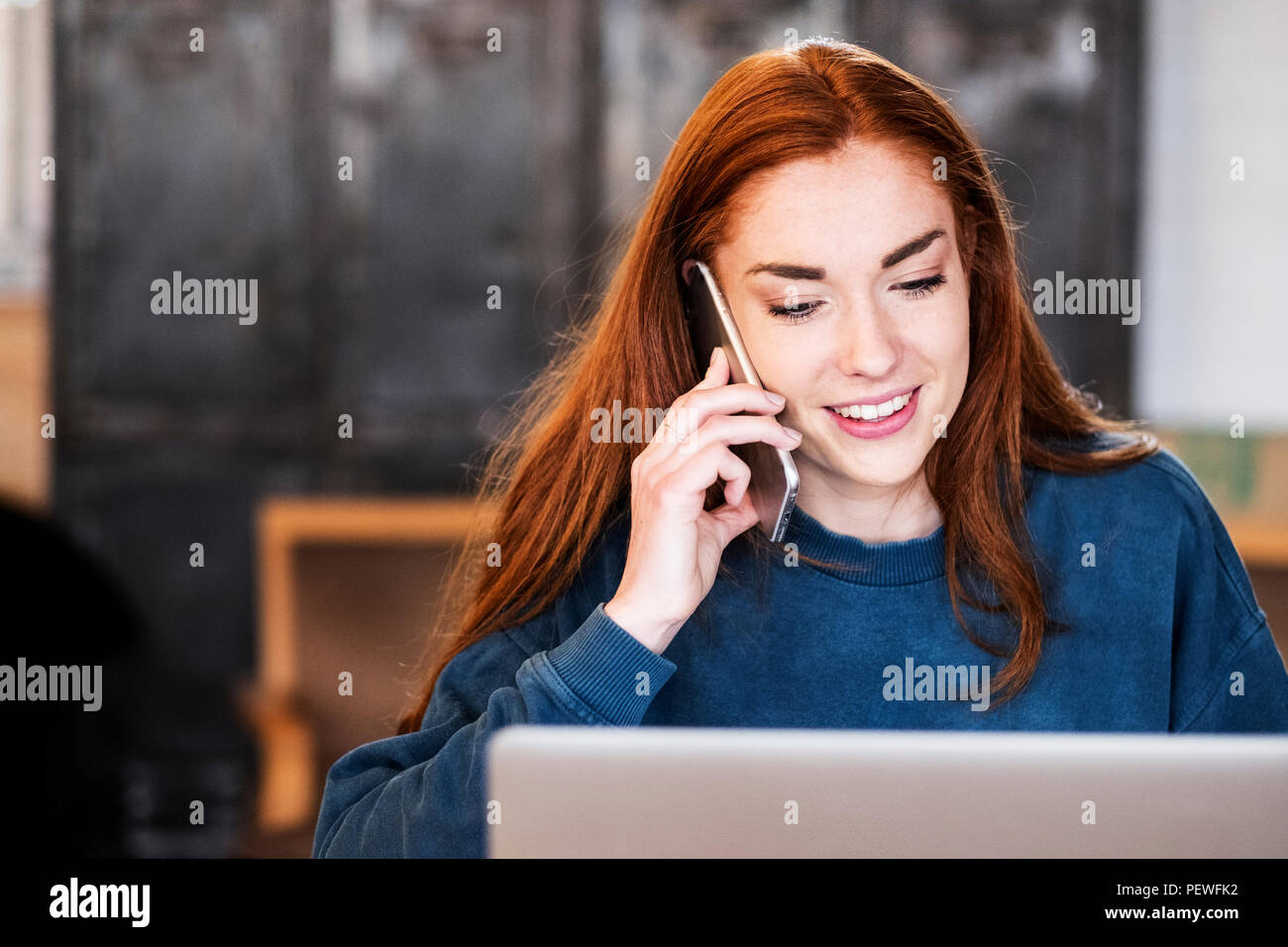 Lächelnde junge Frau mit langen roten Haaren sitzen am Tisch, Arbeiten am Laptop, mit Handy. Stockfoto