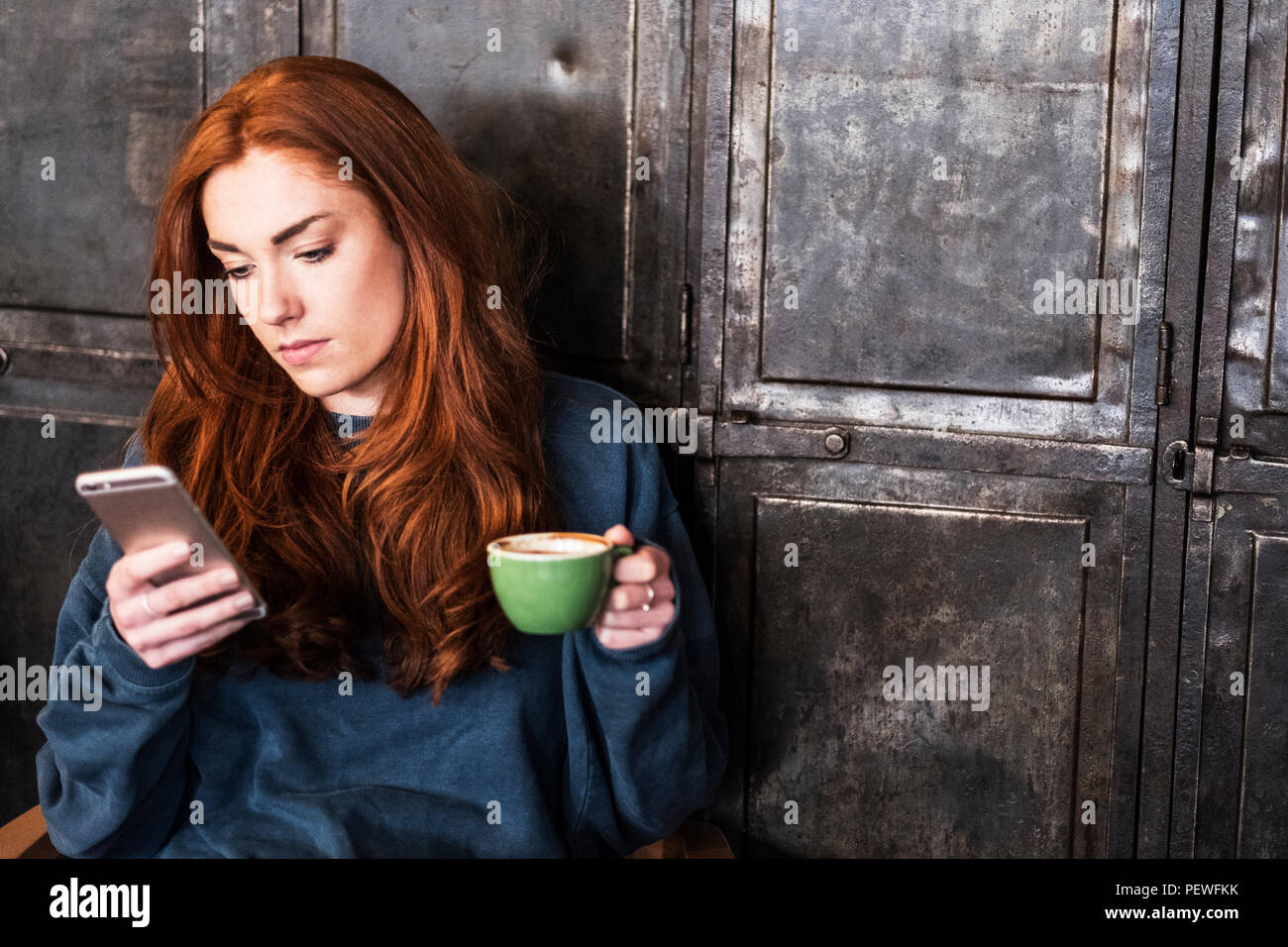 Junge Frau mit langen roten Haaren sitzen am Tisch, Tasse Kaffee, Kontrolle ihr Handy. Stockfoto