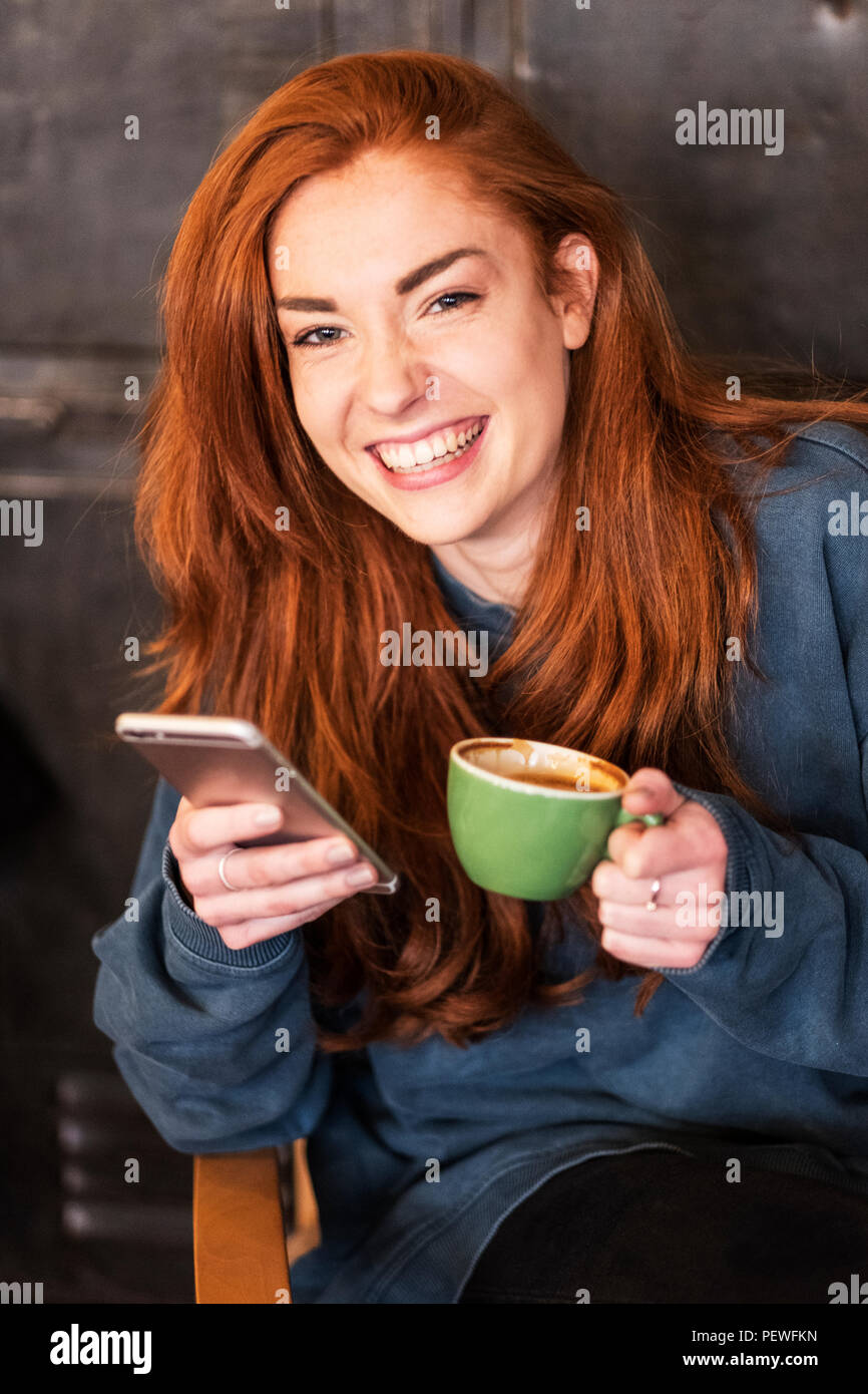 Lächelnde junge Frau mit langen roten Haaren sitzen am Tisch, Handy und Tasse Kaffee, an der Kamera schaut. Stockfoto