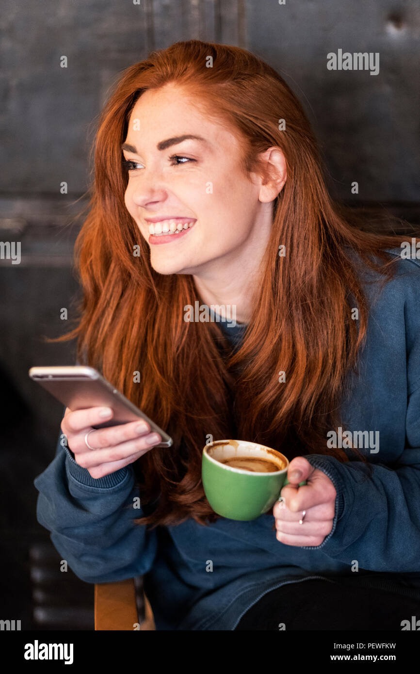 Lächelnde junge Frau mit langen roten Haaren sitzen am Tisch, Handy und Tasse Kaffee. Stockfoto