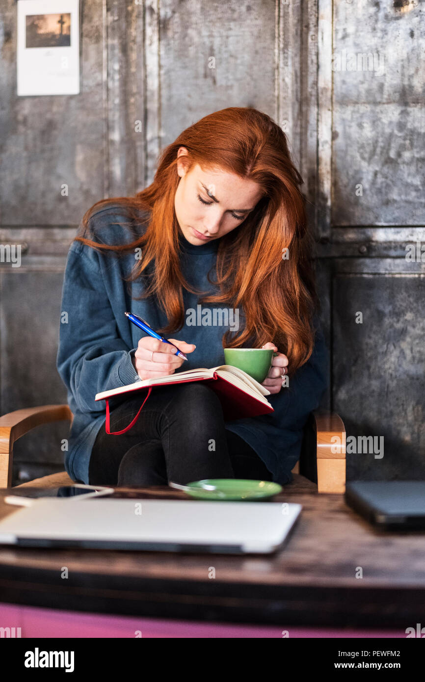 Junge Frau mit langen roten Haaren sitzen am Tisch, Notebook und Tasse Kaffee. Stockfoto