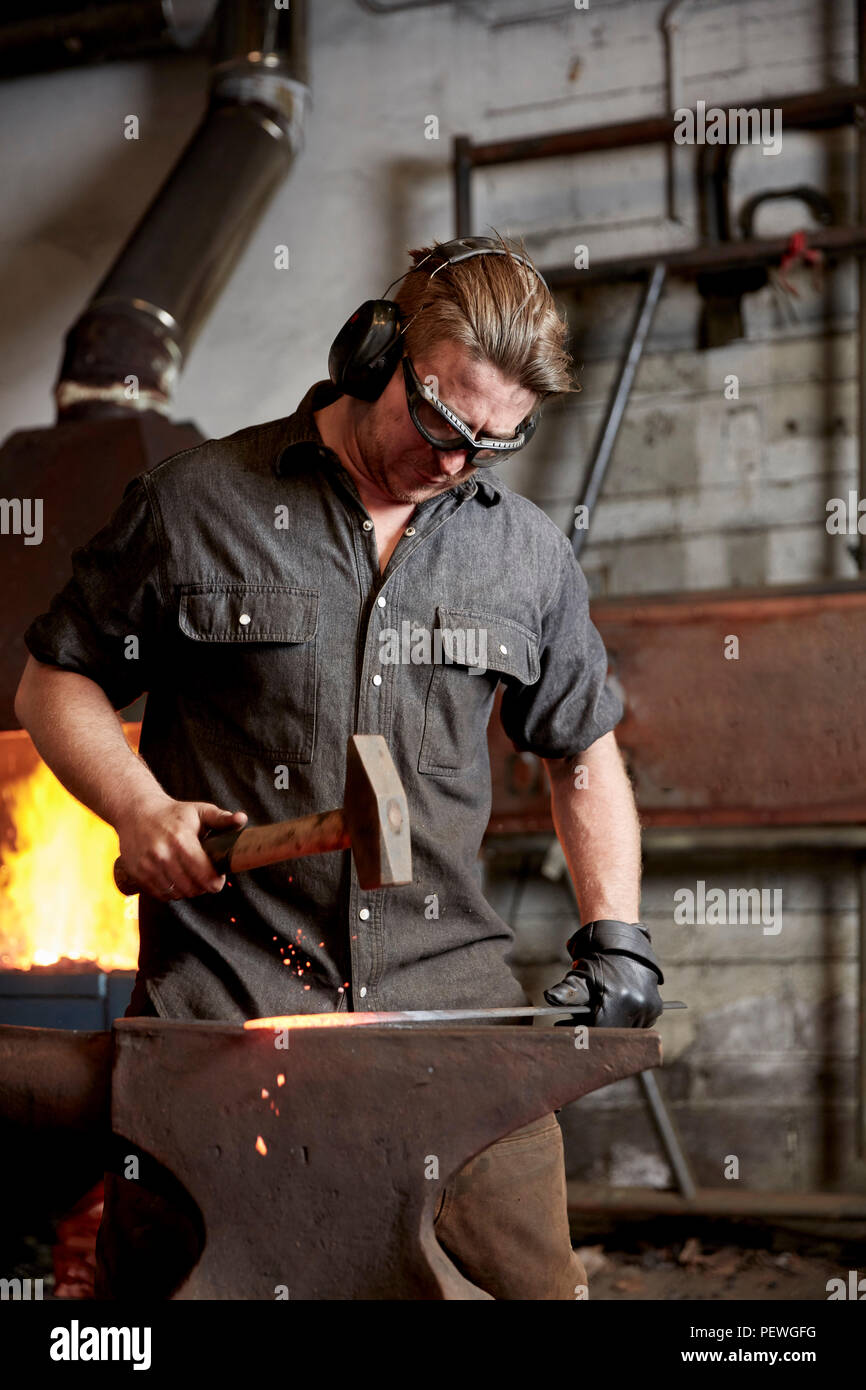 Ein Handwerker Metallarbeiter in Gehörschutz mit einem Hammer ein red hot Stück Metall auf einen Amboss zu gestalten. Stockfoto