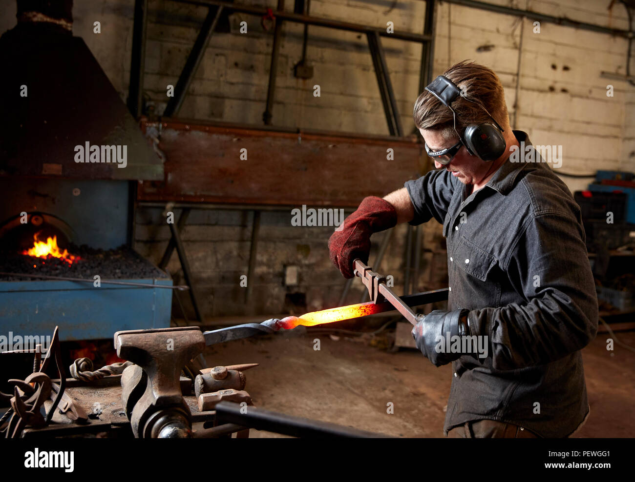 Artisan metall Arbeiter Tragen von Gehörschutz und Schutzbrille mit einem Hammer und Amboss ein red hot Stück Metall mit einem Twist in das Stück zu gestalten. Stockfoto