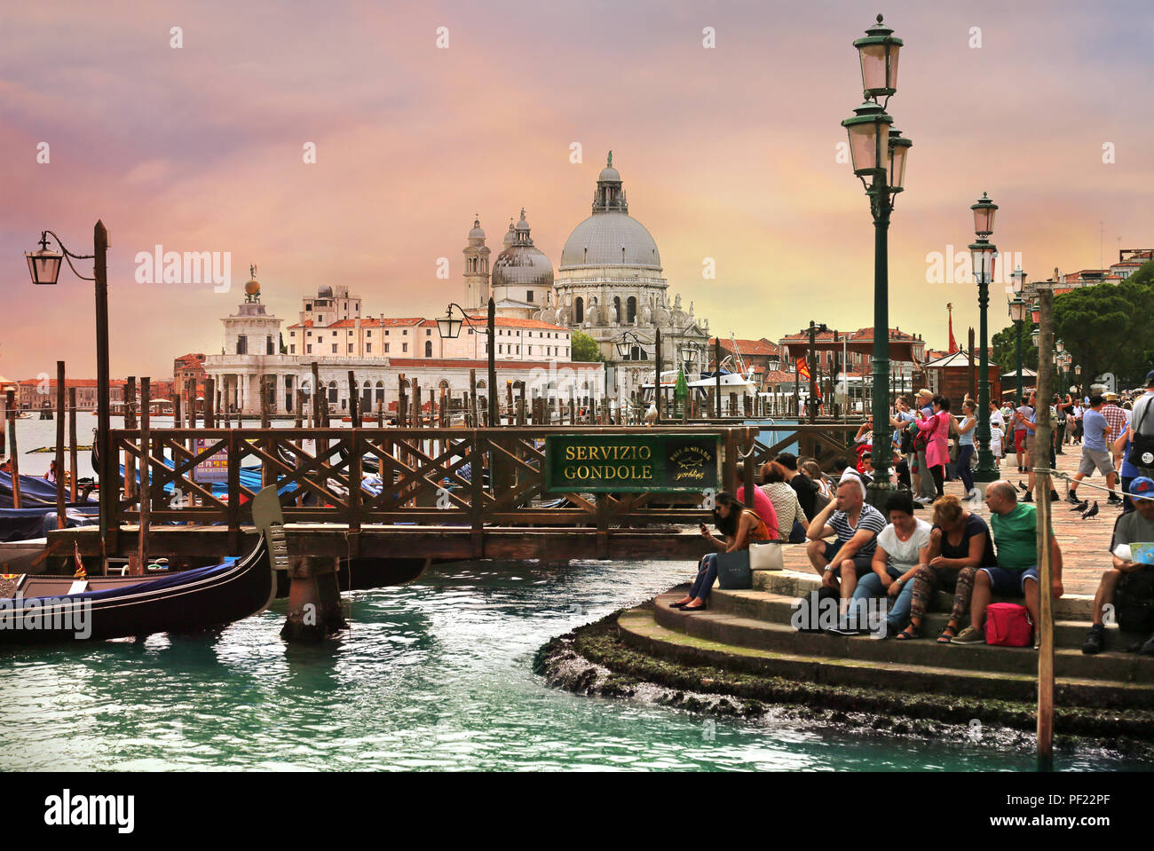 Venedig, Italien, Jun 8, 2018: Touristen sitzen auf die Schritte, die in der Nähe von Gondeln mit Basilika di Santa Maria della Salute im Hintergrund Stockfoto