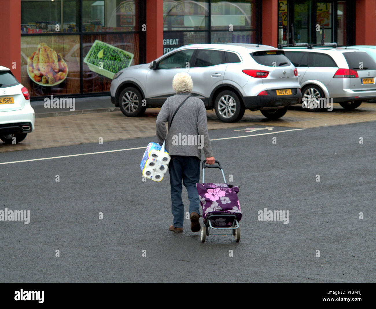 Alte Frau mit Einkaufswagen mit Toilettenpapier Parkplatz in Richtung Fish und Chips Erbsen Anzeigen großer Armut Bereich Drumchapel, Glasgow, Großbritannien Stockfoto