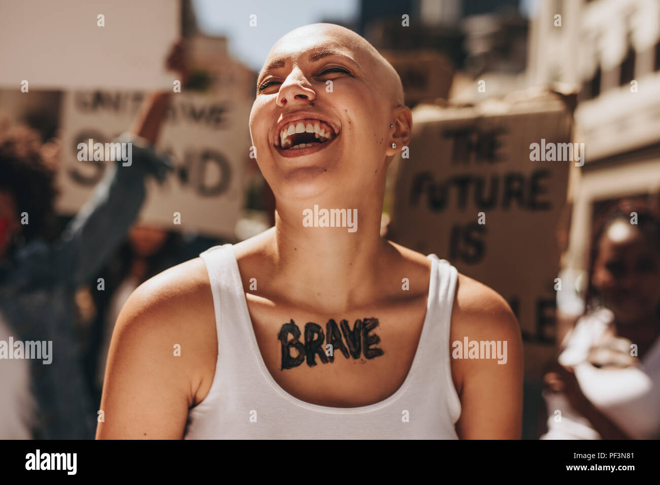 Glatze Frau lachend im Freien während eines Protestes. Mutige Frau mit einer Gruppe von Demonstranten im Hintergrund. Stockfoto