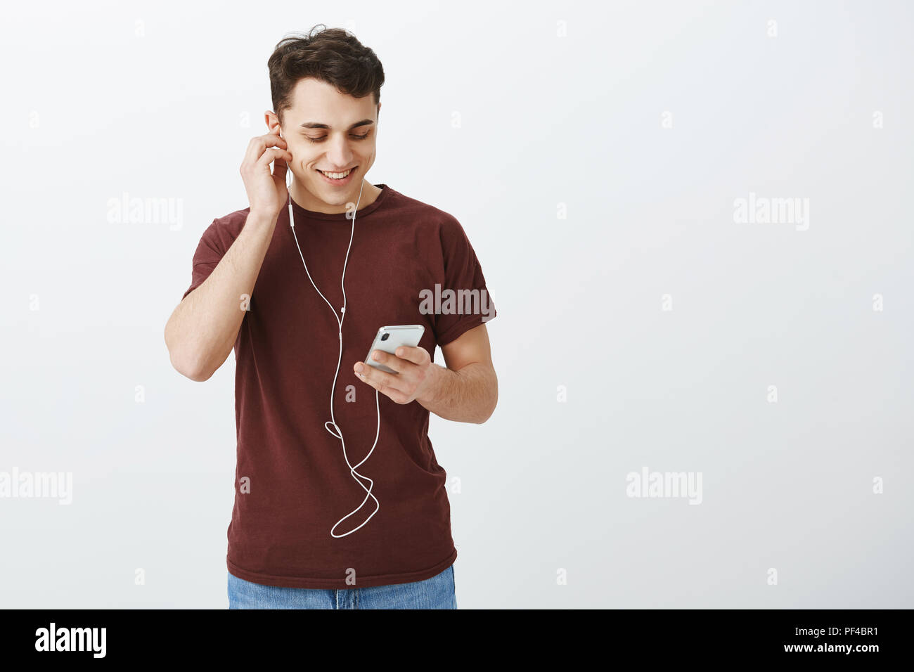 Innen- schuss von zufriedenen Sorglos gut aussehender Kerl in casual rotes T-Shirt, Ohrhörer und breit grinsend am Bildschirm des Smartphones während Pop Musik und genießen Sie hochwertigen Sound Stockfoto