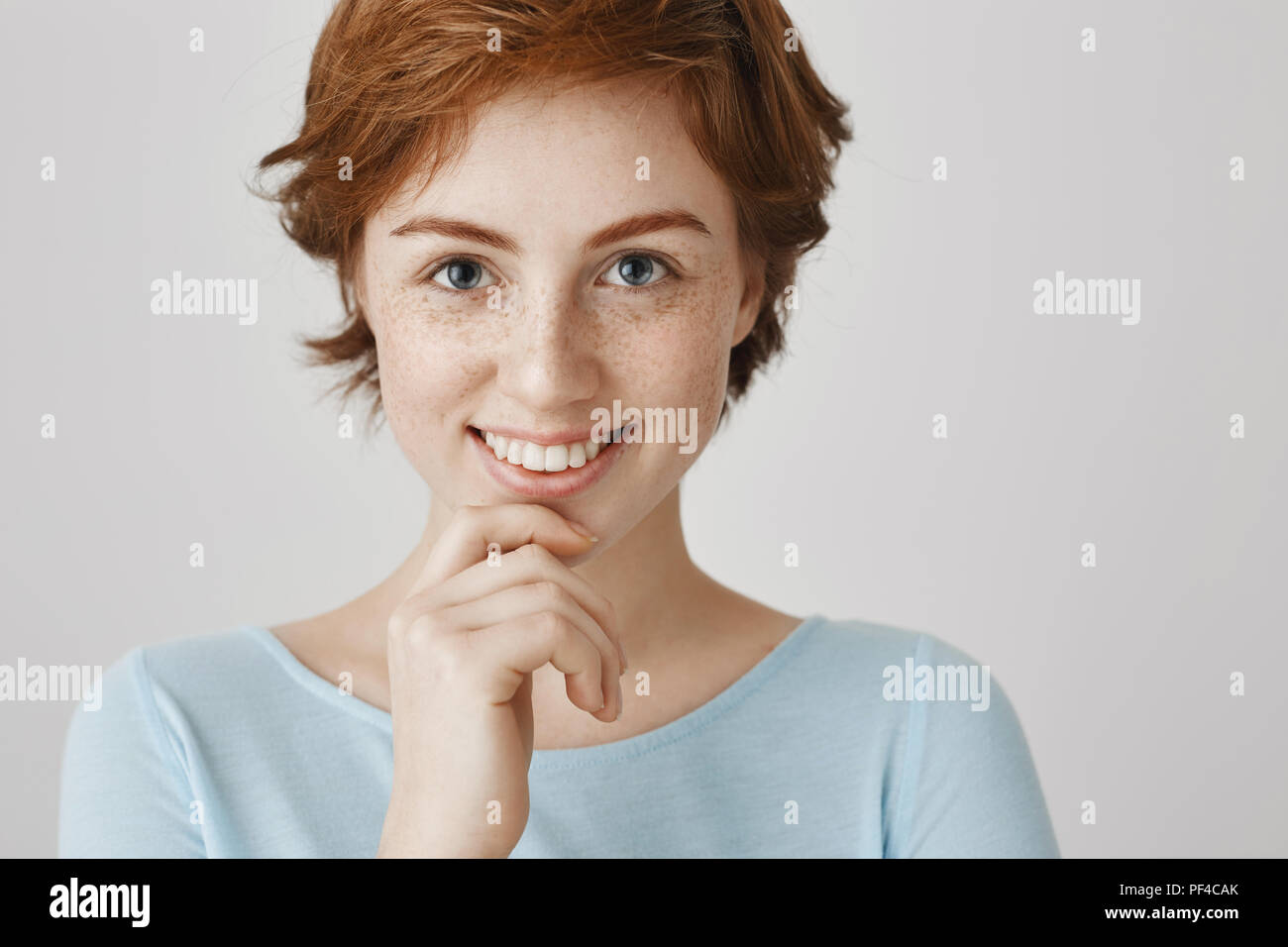 Augen sind die Spiegel der Seele. Close-up Portrait von attraktiven weiblichen mit Ingwer Haar und Sommersprossen die Hand am Kinn und breit grinsend in die Kamera schaut mit interessierten Ausdruck über graue Wand Stockfoto