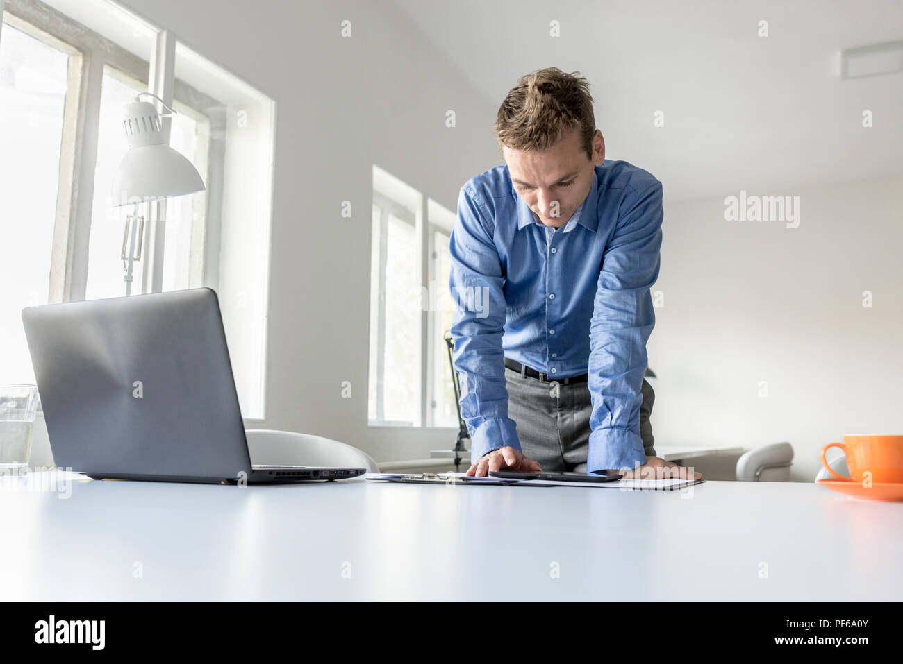 Stilvolle Junge Unternehmer stehend lehnte sich auf seinen Schreibtisch lesen um einen Tablet-PC mit einem ernsten Ausdruck, Vorderansicht. Stockfoto