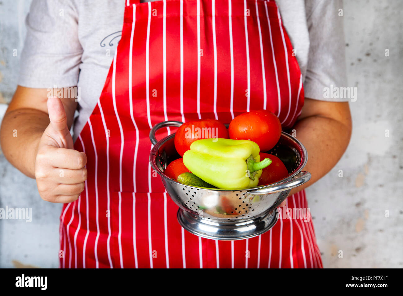Frau in einem roten Schürze mit einem Sieb mit Gemüse für Salat zeigt Hand Zeichen 'ok'. Das Kochen. Stockfoto