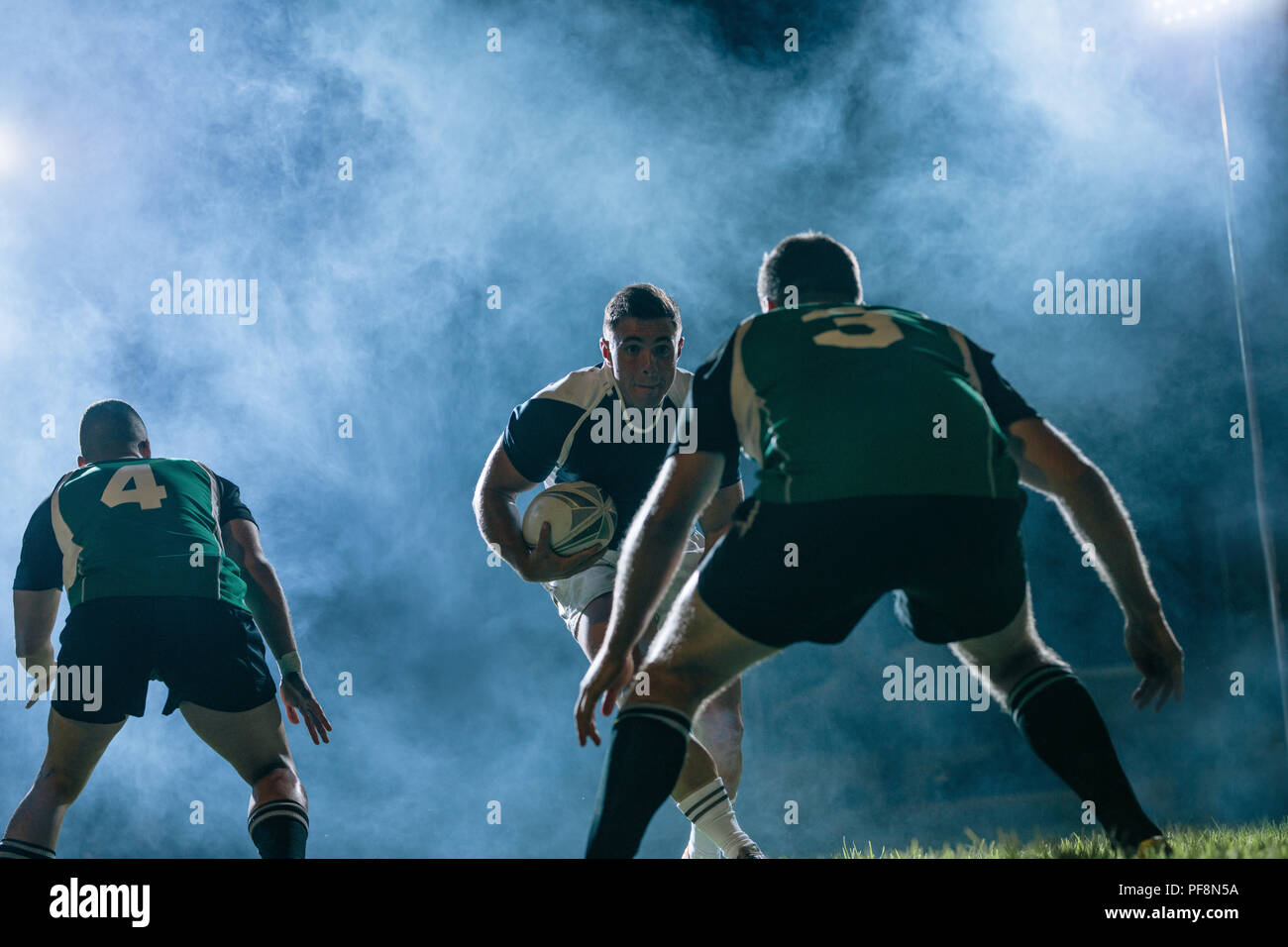 Professionelle rugby spieler läuft mit dem Ball und Gegner team Spieler, die versuchen, ihn auf einer Sport Arena während der Nacht Spiel zu blockieren. Stockfoto