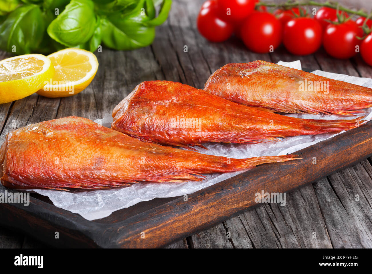 Red Sea Bass auf einem Pergament Papier auf einem alten Holztisch mit Basilikumblätter, Zitronenscheiben und Cherry Tomaten auf den Hintergrund, horizontale close-up Stockfoto