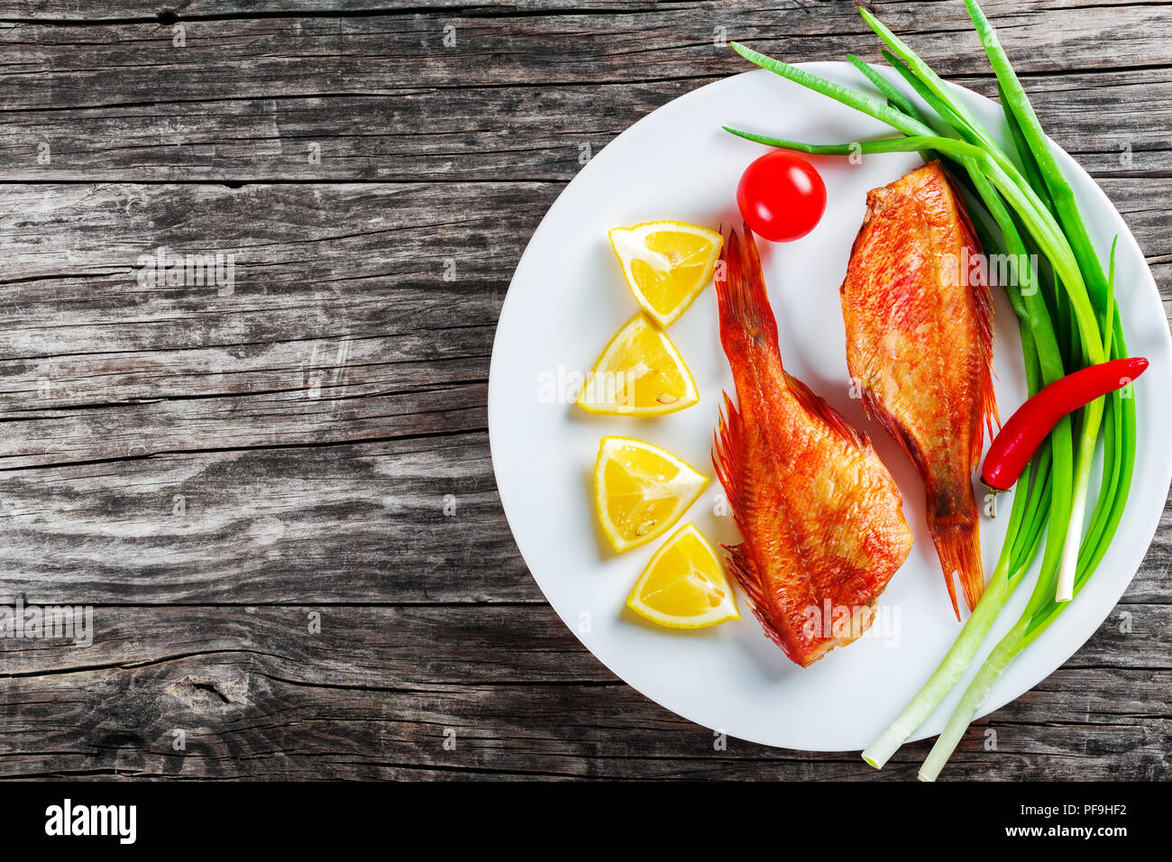 Red Sea Bass auf einem weißen Teller mit grünen Frühlingszwiebeln, Zitronenscheiben und Cherry Tomaten auf einem alten Holztisch, horizontale Ansicht von oben Stockfoto