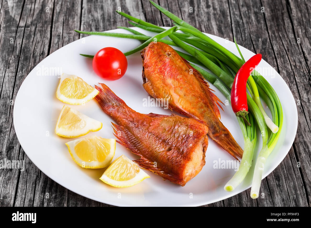 Red Sea Bass auf awhite Teller auf einen alten Holztisch mit grünen Frühlingszwiebeln, Zitronenscheiben und Cherry Tomaten auf den Hintergrund, horizontale close-up Stockfoto