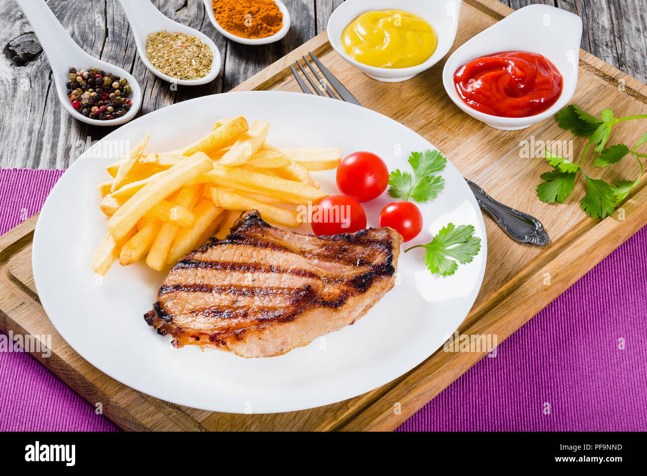 Gegrillte Schweinekoteletts auf einem weißen Teller mit Pommes Frites, Tomaten, Koriander Blätter und Gewürze, auf einem Holztisch, close-up Stockfoto