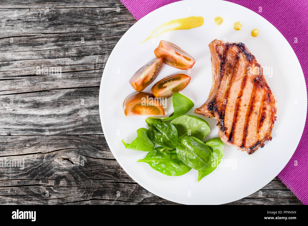 Gegrillte Schweinekoteletts auf einem weißen Teller mit Scheiben von schwarzen Tomaten, Spinat auf einen Tisch Matte auf einem hölzernen tabl Blätter, Ansicht von oben Stockfoto