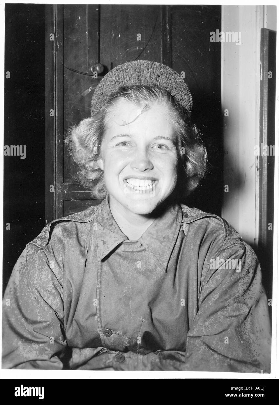 Schwarz-weiß Foto zeigt einen lächelnden jungen blonden Frau, von der Taille bis, das Tragen eines verregneten militärischen Jacke und eine Mütze mit Krempe, sitzt vor einem Schaltschrank, wahrscheinlich fotografierte während des Zweiten Weltkrieges, 1945. () Stockfoto